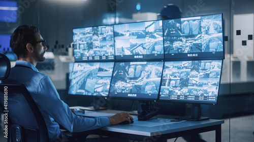 Industry 4.0 Modern Factory: Security Operator Controls Proper Functioning of Workshop Production Line, Uses Computer with Screens Showing Surveillance Camera Feed. High-Tech Security