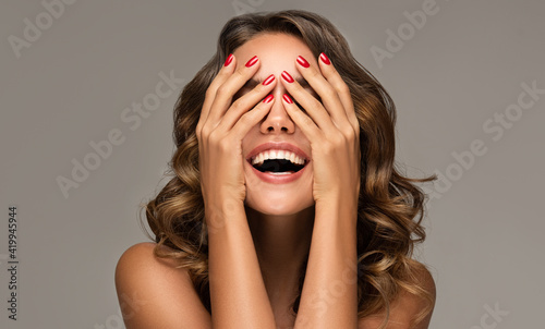 Beautiful woman with curly hair and red nails manicure . Girl happy laughs closes her face with a hand .