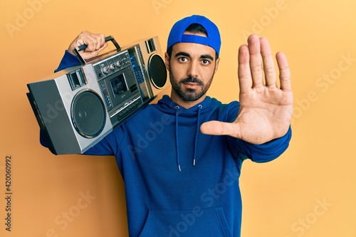 Young hispanic man holding boombox, listening to music with open hand doing stop sign with serious and confident expression, defense gesture