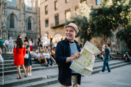 Happy Caucasian man 60s enjoying solo trip vacations for exploring Italy during retirement, cheerful male tourist in straw hat using paper orientation map for walking around historis center