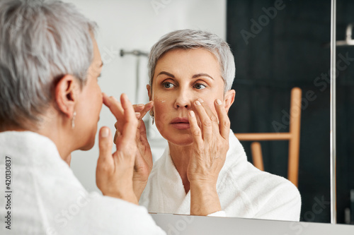 Mature woman applying moisturizer in bathroom. Senior female in bathrobe doing skincare routine.