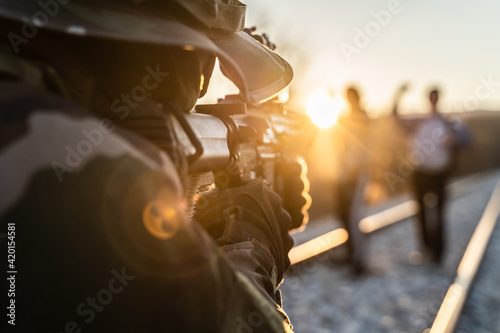Group of two migrants or terrorists held by soldier at border patrol on the railroad in day -Two unknown men raised arms facing armed man in uniform on road after being arrested for illegal activity