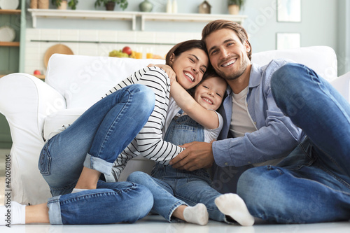 Young Caucasian family with small daughter pose relax on floor in living room, smiling little girl kid hug embrace parents, show love and gratitude, rest at home together.