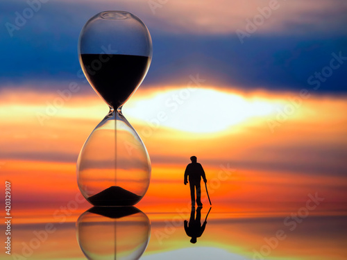 miniature people. silhouette of an elderly man walking towards the sunset next to an hourglass. end of life.