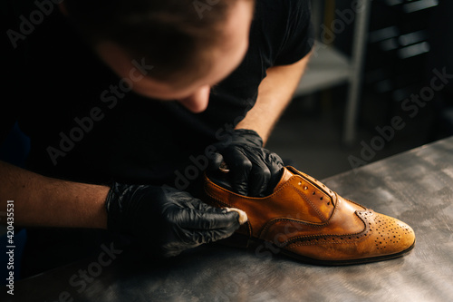 Top close-up view of professional shoemaker wearing black gloves polishing old light brown leather shoes. Concept of cobbler artisan repairing and restoration work in shoe repair shop.