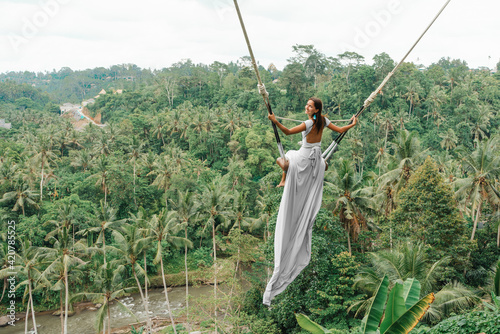 Tanned beautiful woman in a long white dress with a train, riding on a swing. In the background, a rainforest and palm trees. Copy space. Rear view