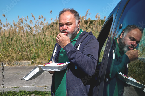 Funny bearded mature man eating vegan sandwich on the street near the car. Unhealthy eating. Fast food. Takeaway food