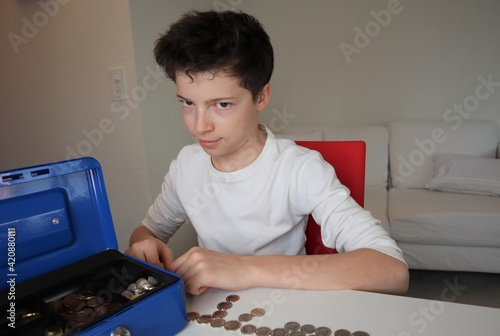 Boy with light skin and brown hair secretly counting the money and coins that he has saved in his lock box. Rows of coins are on the table.