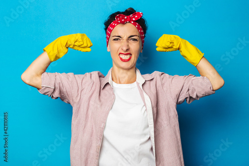 Strong young woman showing biceps in cleaning gloves on blue background.