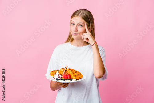 Young russian woman eating a waffle isolated pointing temple with finger, thinking, focused on a task.