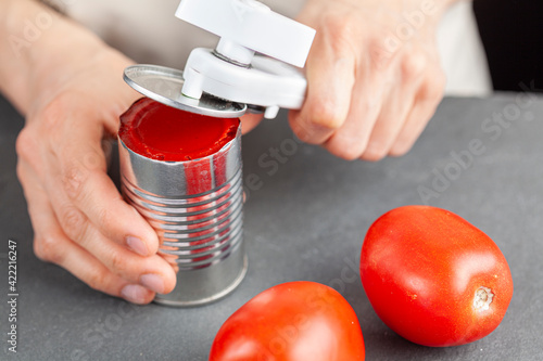 A woman is carefully opening a can of tomato paste on a kitchen counter using a white plastic can opener. She is preparing a meal for which she uses both fresh and pureed preserved tomatoes