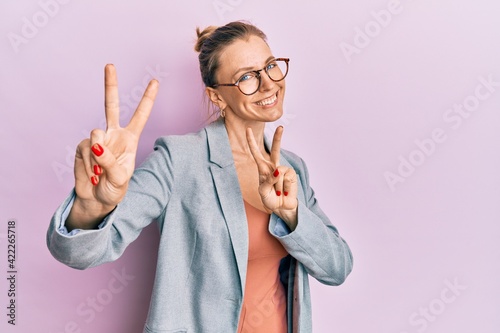 Beautiful caucasian woman wearing business jacket and glasses smiling looking to the camera showing fingers doing victory sign. number two.