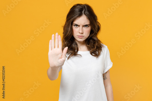 Young serious strict angry eueropean caucasian student woman 20s in white basic casual blank print design t-shirt show stop palm gesture refusing isolated on yellow orange background studio portrait.