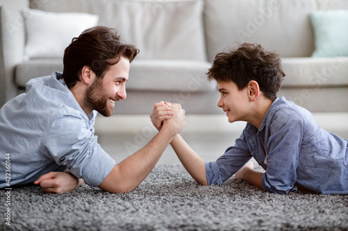 Happy father competing in arm-wrestling with his son, enjoying time together at home, side view