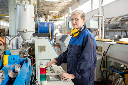 Young female worker in overalls looking at you while standing in front of control panel of industrial machine and preparing it for work