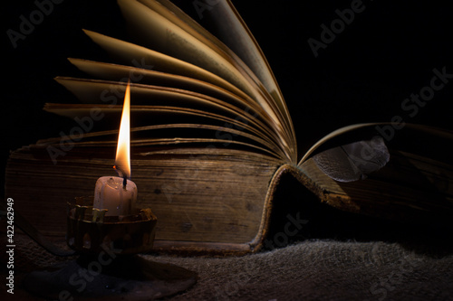 A candle burns in the dark. In the background there is an old book with open pages. The concept of mysticism and secret knowledge, esotericism. Selective focus on a burning candle.