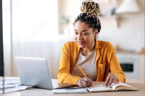 Focused cute stylish african american female student with afro dreadlocks, studying remotely from home, using a laptop, taking notes on notepad during online lesson, e-learning concept, smiling