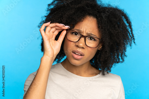 Studio portrait of unsure doubtful African American girl touching glasses, suspicious looking at camera, isolated on blue blank background. Sceptical mistrustful Black woman adjusts eyeglasses