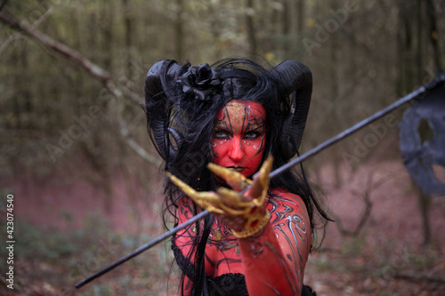 Sexy Teufelin steht im Wald. Sie hat die Hörner auf dem Kopf, eine schwarze Corsage. sie ist mit der roten Farbe und der Muster angemalt.Sie hat lange, schwarze Haare.Überall ist der roter Rauch.