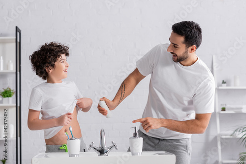 Smiling arabian man holding deodorant near son and sink in bathroom