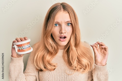 Beautiful young caucasian girl holding invisible aligner orthodontic and braces afraid and shocked with surprise and amazed expression, fear and excited face.
