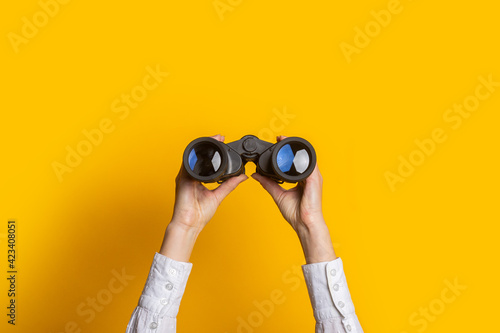 female hands hold black binoculars on a bright yellow background