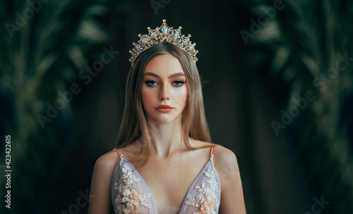 Portrait of fantasy medieval girl princess in dark gothic room. Woman queen looking at camera, beauty face. Vintage trendy glamour dress golden luxury crown, long loose blonde hair. Fashion model