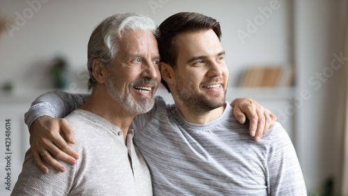 Smiling young Caucasian man and older father hug look in window distance dreaming imagining together. Optimistic mature dad and adult grownup son embrace visualize happy future. Vision concept.