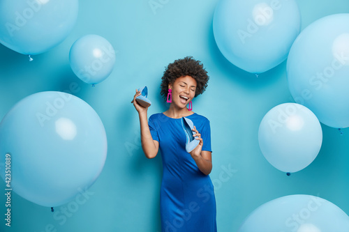 Positive African American woman with curly hair keeps shoes near mouth pretends singing wears dress chooses outfit for party event isolated over blue background with inflated balloons around