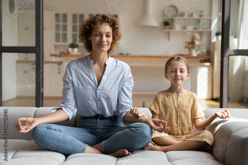 Calm young Caucasian mother and little 9s daughter sit relax on sofa in living room practice yoga together. Happy mom and small girl child rest meditate on couch at home, relieve negative emotions.