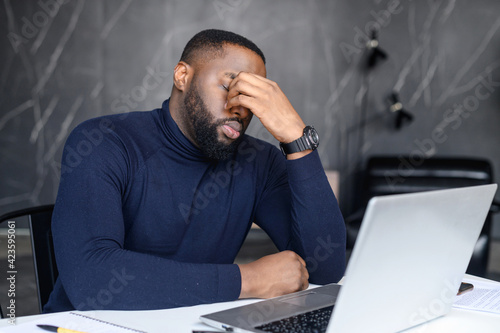 Difficult task from customer. African American man conducts correspondence using a laptop, hearing bad unexpected news, scared young male holds his hand near the bridge of the nose, sitting at desk