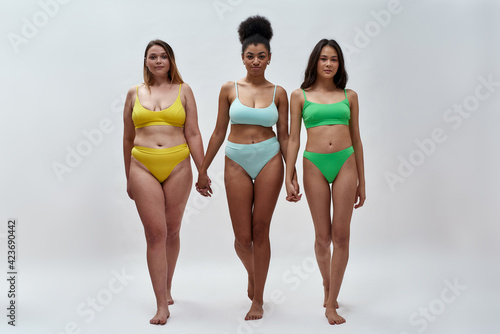 Full length shot of three confident diverse women in colorful underwear holding hands, looking at camera, standing together isolated over light background