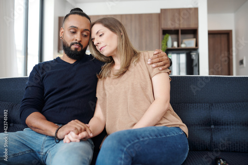 Sad young mixed-race couple reassuring each other when sitting on sofa at home