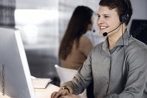 Young dark-haired guy in a green shirt and headsets is talking to a client, while sitting at the desk, working together with a female colleague in a modern office. Call center operators at work