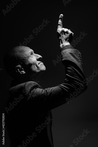 Black and white portrait of a brutal middle-aged bald man. A charismatic man, illuminated by a spotlight in a black suit against a black background, looks up and points up with his hand.
