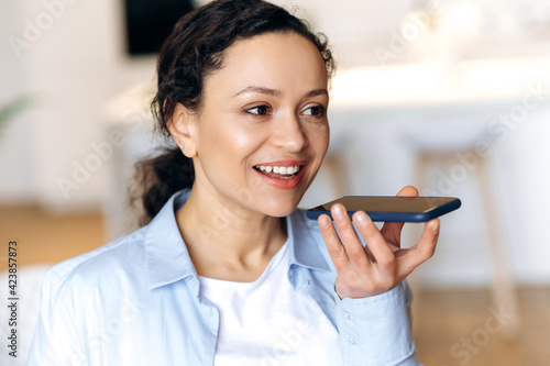 Attractive successful mixed race brunette attractive woman, using smartphone, talking to friends or colleagues on speakerphone or recording audio message, looking away, smiling friendly