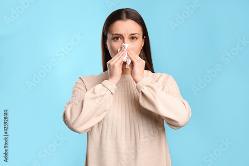 Young woman with tissue suffering from runny nose on light blue background