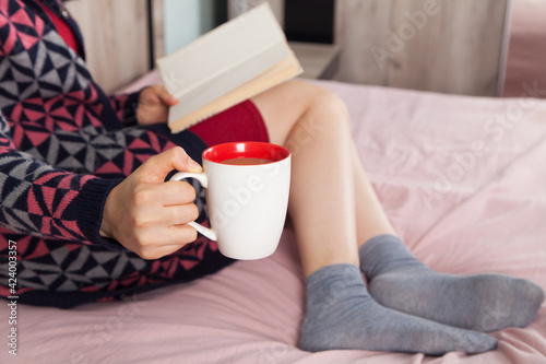 Young woman is relaxing in her bed with coffee and book. She is wearing a socks.