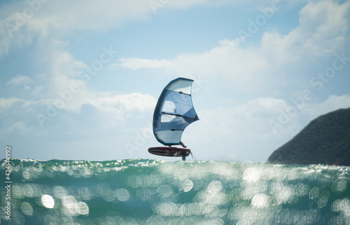 A man wing foils at sea using a hand held inflatable wing, riding a hydrofoil surfboard. Turquoise sea, cloudy blue sky. 