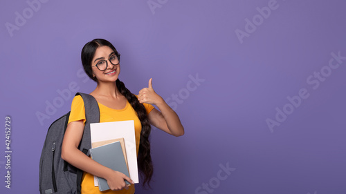 Smiling indian student wearing eyeglasses holding notebooks showing thumbs up