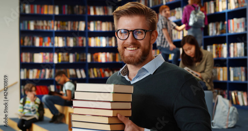 Bearded school teacher or librarian holding books in hands standing in library interior.