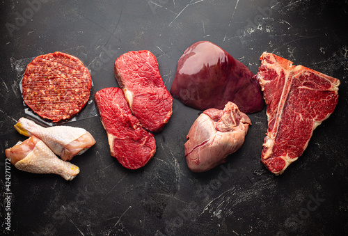 Selection of assorted raw meat food for zero carb carnivore diet: uncooked beef steak, ground meat patty, heart, liver and chicken legs on black stone background from above 