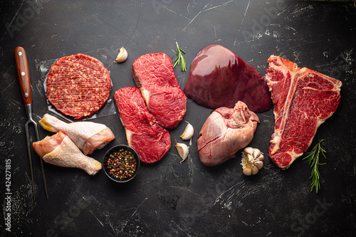 Selection of assorted raw meat food with seasonings for zero carb carnivore diet: uncooked beef steak, ground meat patty, heart, liver and chicken legs on black stone background from above 