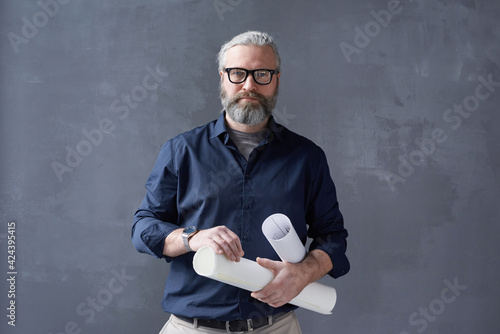 Portrait of mature bearded architect in eyeglasses standing with blueprints against the black background