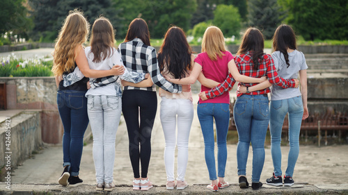 Seven athletic volleyball girls with beautiful priests of fit young slim girls in shirts and jeans hugging each other look into the distance with their backs to the camera. 