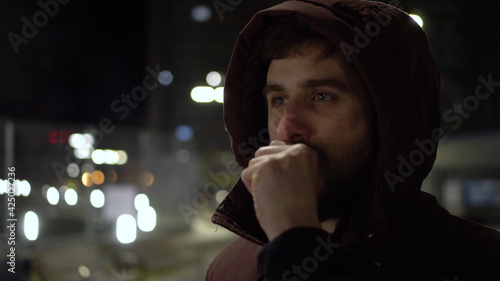 A young man stands in the cold in the evening outside and warms his hands with his breath.