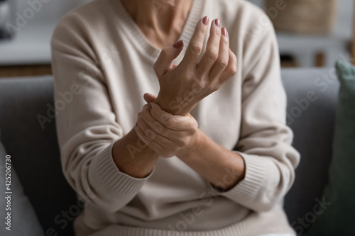 Middle aged elderly woman feeling wrist pain, sitting on couch at home, holding arm. Senior 60s pensioner suffering from rheumatoid arthritis, joints ache, muscle strain, inflammation. Close up