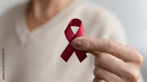 Lady showing hiv or aids awareness symbol, volunteering for charity campaign for prevention immunity disease, cancer, elderly healthcare support. Hand of mature woman holding red ribbon. Close up