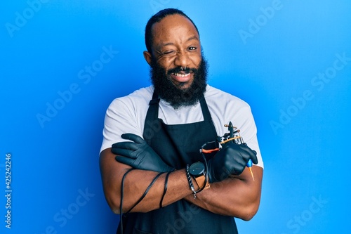 Young african american man tattoo artist wearing professional uniform and gloves holding tattooer machine winking looking at the camera with sexy expression, cheerful and happy face.