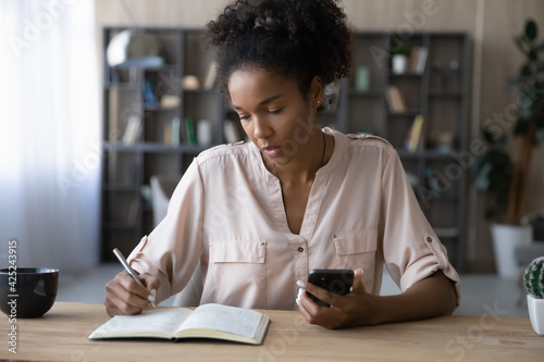 Focused young African American woman make notes handwrite in notebook use application on smartphone. Millennial biracial female write make list plan with cellphone. Planner, management concept.